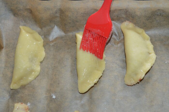 Unbaked crescents arranged on a parchment-lined tray being brushed with egg white